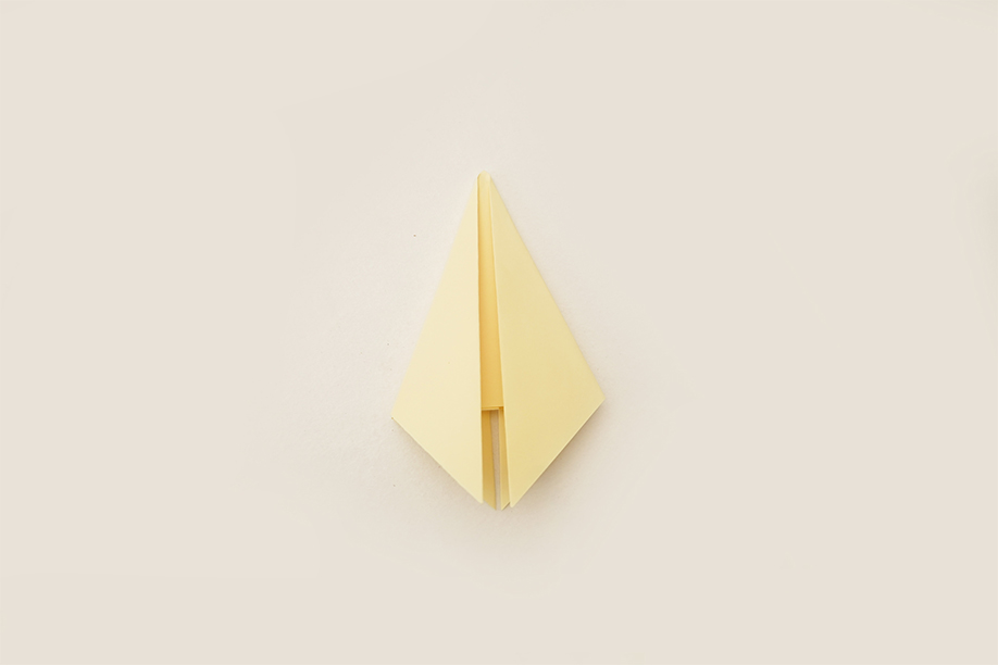 A pale yellow square of paper, following previous folds, is now folded into a more complex, symmetrical shape. The paper is folded in half, creating a central crease, then the top and bottom points are folded inwards towards the centre crease. The resulting shape is roughly diamond-shaped, with a central vertical slit. The piece lies flat on a plain white background, the even lighting clearly showing the precise folds and uniform colour of the paper. The image depicts another step in a paper-folding process, likely origami, progressing from previous stages.