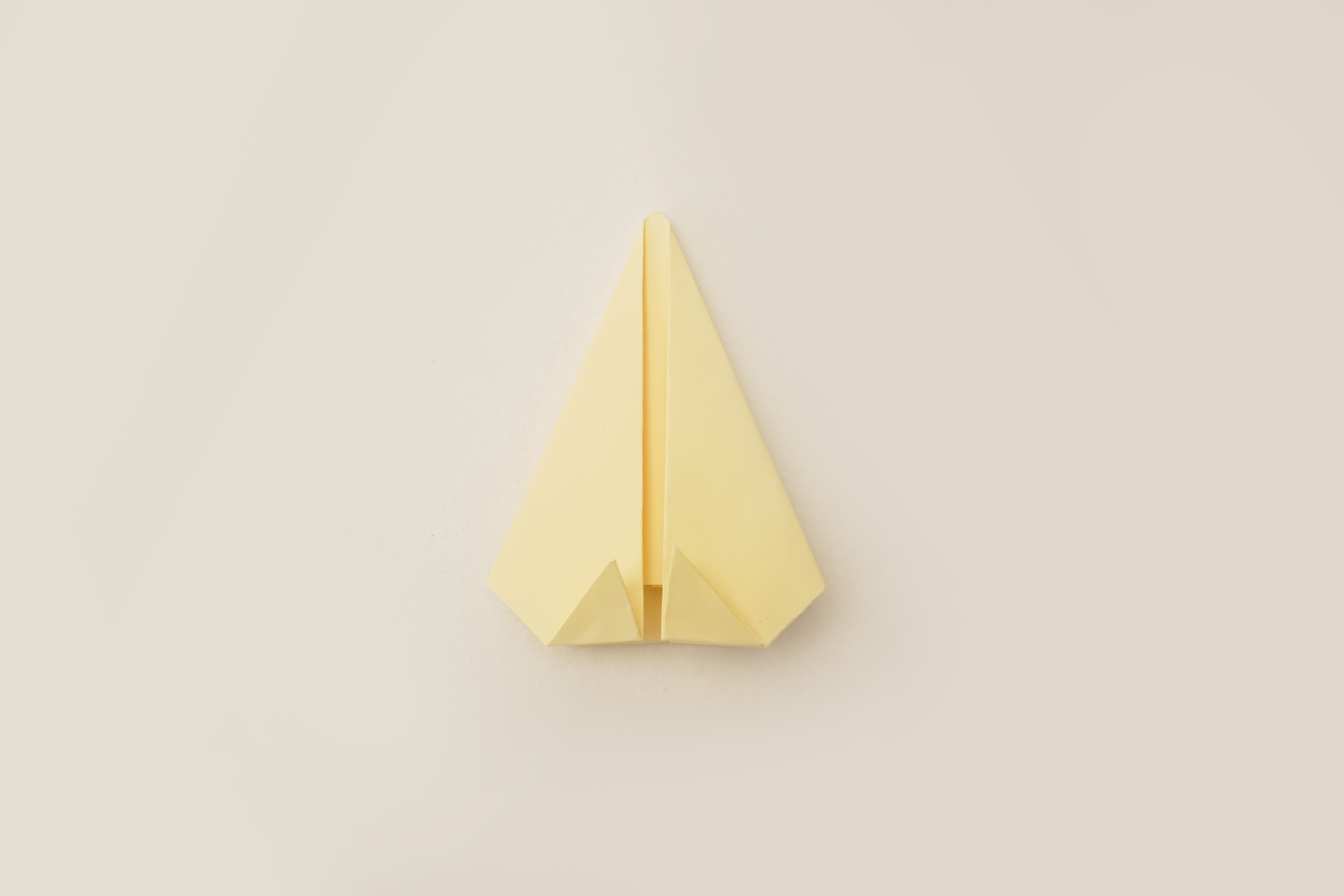 A pale yellow square of paper, following previous folds, is now shaped like a simple, upright isosceles triangle. The base of the triangle is slightly wider than the top, and the overall form is symmetrical. The paper is neatly folded, with sharp creases, and lies flat against a plain, off-white background. The even lighting clearly shows the paper's uniform colour and the precision of the folds. The image shows another stage in an origami project, building upon previous steps.