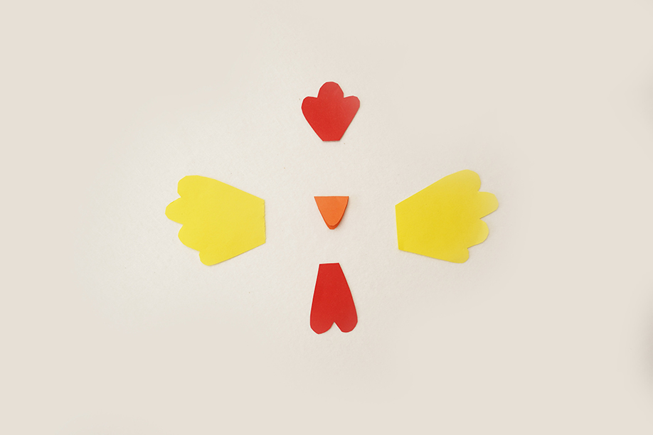Five brightly coloured pieces of card are arranged on a plain white background. Two yellow shapes, representing wings, are positioned symmetrically on either side of a small, orange triangle, which forms the chicken's beak. Above the beak is a red shape, representing the comb, and below is another red shape, forming the wattle. The shapes are simple, almost cartoonish, and neatly positioned, suggesting preparation for a craft project. The overall impression is one of a playful and straightforward craft design.