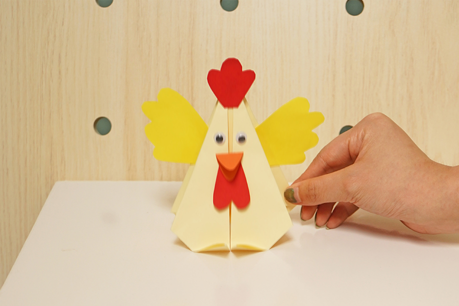 A hand gently adjusts a completed origami chicken, which stands on a white surface against a light wood-panelled background. The chicken is predominantly cream-coloured, with bright yellow wings and red detailing for its comb and wattle. Simple, round, black googly eyes add to its charm. The overall impression is one of a successfully completed craft project, showcasing a simple yet effective origami design. The image is bright, clean, and focuses on the finished model.