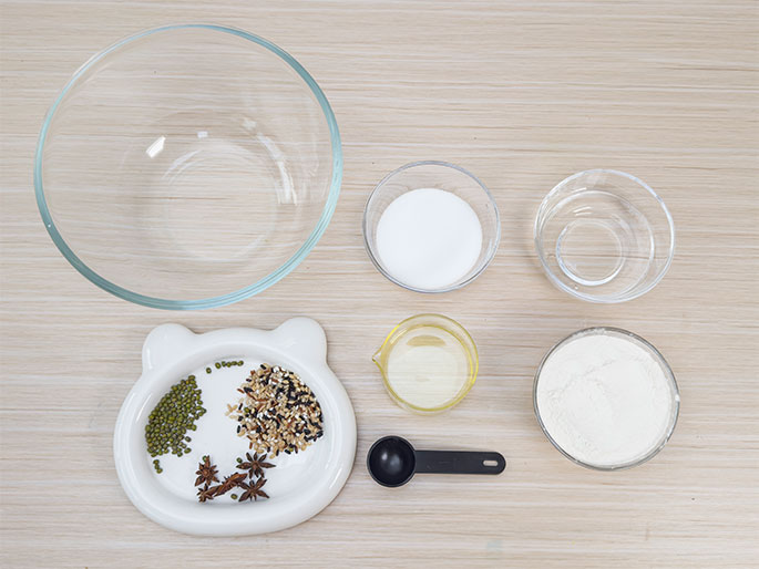 An assortment of ingredients is laid out on a light wood-grained surface, suggesting preparation for a recipe. A large, empty clear glass bowl sits to the left. Smaller clear glass bowls containing sugar, water, and flour are arranged to the right. A small yellow container holds oil, and a black measuring spoon lies nearby. A white, cat-shaped dish contains star anise, mung beans, and a mix of rice and sesame seeds. This image depicts the mise en place for a cooking or baking project.