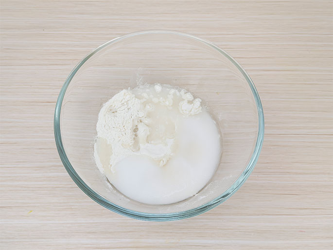 A clear glass bowl, placed on a light wood-grained surface, contains the initial stages of a mixture. White flour, sugar, and a clear liquid, likely water, are visible but not yet fully combined. The ingredients appear layered, with the flour mostly on one side and the sugar dissolved into the water. This image represents the beginning stages of a recipe, before mixing.