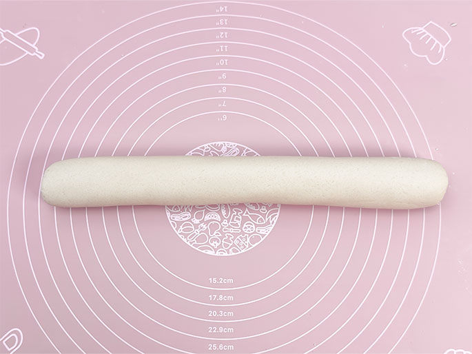 A long, cylindrical roll of pale dough rests on a pink silicone baking mat. The mat features concentric circles with size markings in centimeters and illustrations of food items like a chef's hat, a rolling pin, and various fruits and vegetables. The dough appears smooth and evenly rolled. This image likely depicts a stage in baking or cooking, after the dough has been kneaded and shaped.