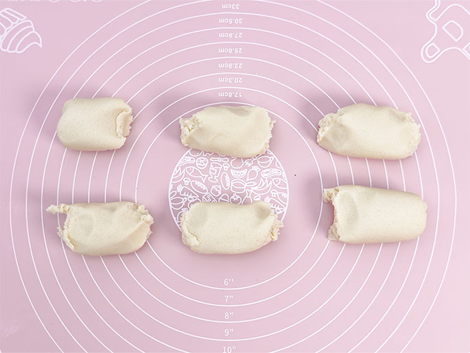 Six roughly equal portions of pale dough are arranged on a pink silicone baking mat. They are oblong in shape, somewhat resembling thick, short logs. The mat has concentric circles marked with measurements in both centimeters and inches, along with illustrations of food-related items. This image shows a step in a baking process where the dough has been divided in preparation for shaping individual pieces.