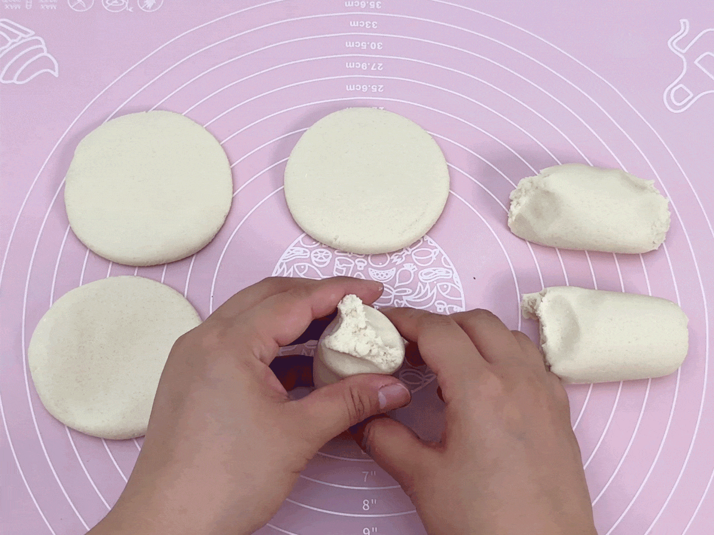 Two hands are working with portions of pale dough on a pink silicone baking mat. Two pieces of dough have been flattened into circles, while two more oblong pieces remain unshaped. The hands are in the process of shaping another piece, pinching it into a thicker form. The mat features concentric circles with measurement markings in inches and centimeters, and illustrations of food-related objects. This image captures a moment in the preparation of baked goods, where the dough portions are being shaped.