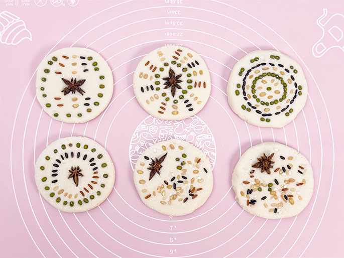 Six round pieces of dough are arranged on a pink silicone baking mat. Each piece is decorated with a variety of small seeds and grains, and a star anise in the center. The seeds and grains vary in color and type, creating different patterns on each dough circle. The mat has concentric circles with measurement markings and food-related illustrations. These appear to be decorative flatbreads or cookies ready for baking.