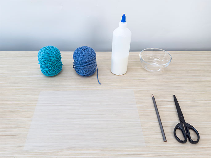 Craft supplies are arranged on a light wood table. Two balls of yarn, one teal and one darker blue, sit next to a bottle of white glue and a small clear glass bowl. A sheet of clear plastic lies in the center of the table, with a pencil and a pair of black scissors placed to its right. These materials appear to be set up for a yarn-based craft project.