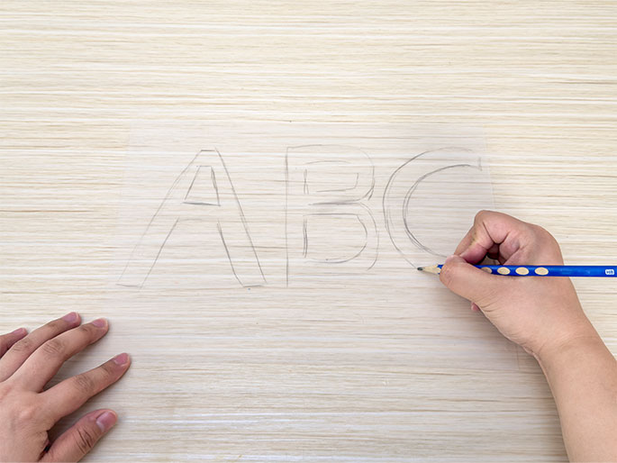 A hand holding a pencil is drawing the letters ABC in block style onto a clear plastic sheet. The sheet rests on a light-colored wooden table. Another hand is visible at the left edge of the frame, holding the plastic sheet in place. The letters are lightly sketched. This image shows the process of creating a template for a craft project.