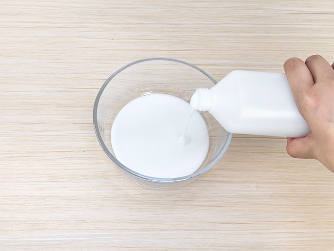 A hand pours white glue from a plastic bottle into a clear glass bowl. The bowl sits on a light wood table. A stream of glue is visible flowing from the bottle into the bowl. This image shows the preparation of glue for a craft project.