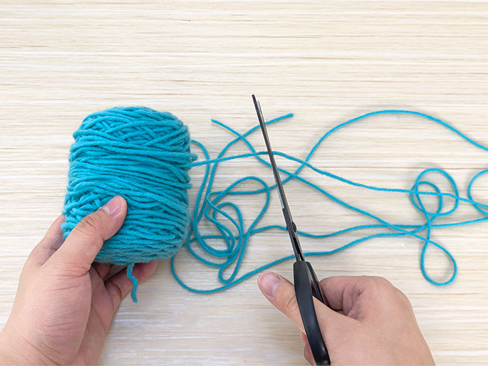 Two hands are engaged in a crafting activity on a light wood surface. One hand holds a ball of teal yarn, while the other hand uses black scissors to cut lengths of the same yarn. Several strands of cut yarn lie on the table. This image demonstrates the process of cutting yarn for a craft project.