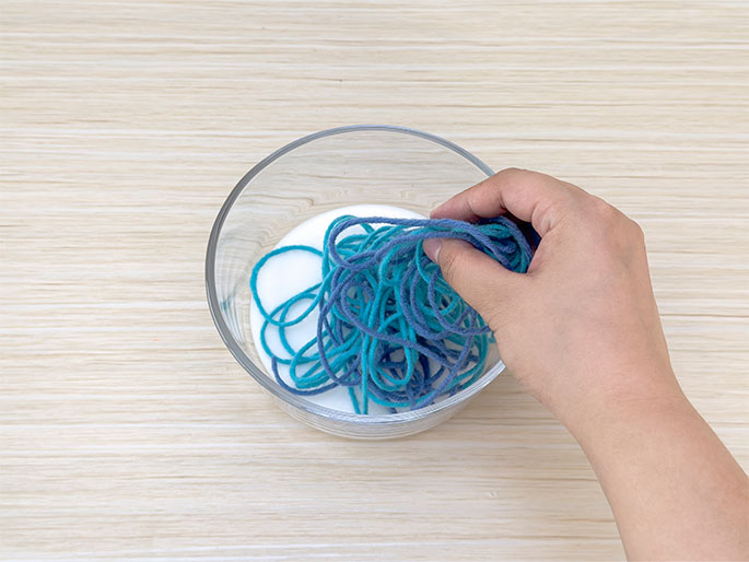 A hand places strands of teal and blue yarn into a clear glass bowl containing white glue. The bowl sits on a light wood table. The yarn is being mixed with the glue, likely in preparation for a craft project.