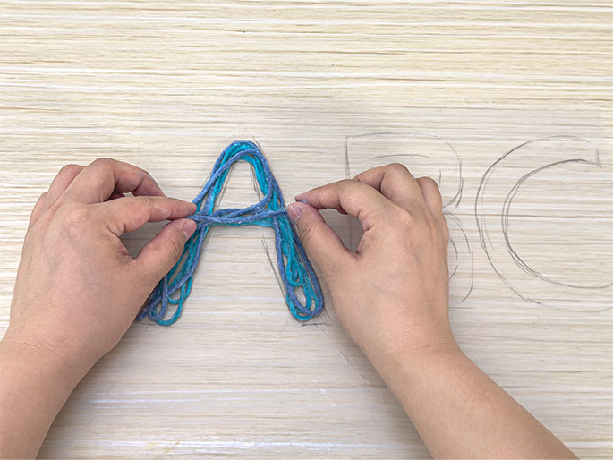 Two hands are arranging teal and blue yarn over a lightly drawn letter A on a clear plastic sheet. The sheet rests on a light wood table. Faint outlines of the letters BC are also visible on the sheet to the right of the A. The yarn appears to be glued down, following the pencil outline of the letter. This image shows the process of creating yarn letters.