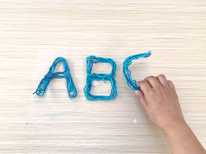 A hand is completing the letter C formed from teal and blue yarn glued onto a clear plastic sheet. The completed yarn letters A and B are also visible on the sheet, which rests on a light wood table. This image shows the process of creating yarn letters.