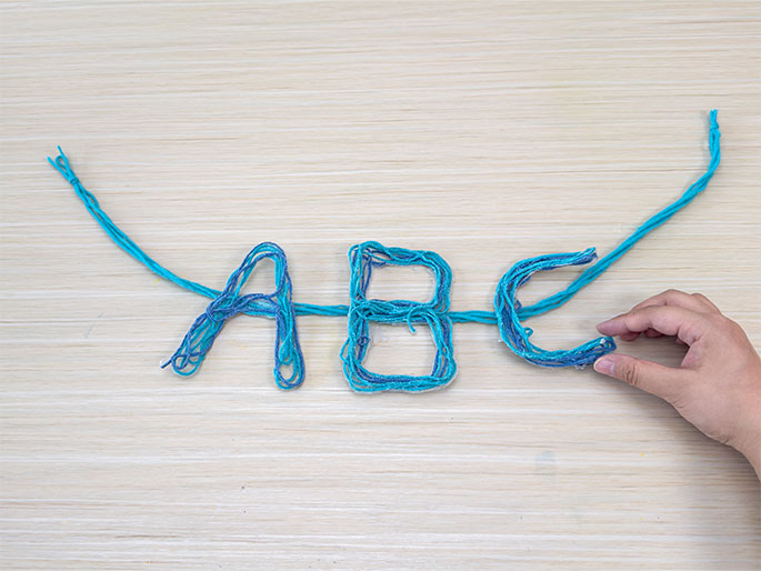 A hand adjusts the yarn letters ABC created from teal and blue yarn glued to a single strand of teal yarn. The letters rest on a light wood table. This image shows completed yarn letters ready to be used for decoration or part of a larger craft project.