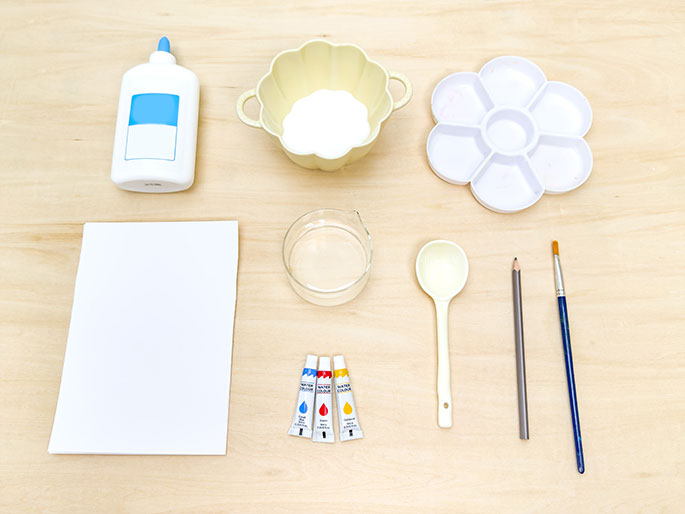 An assortment of art supplies is arranged on a light wood table. The supplies include a bottle of white glue, a pale yellow bowl containing white powder (likely salt or sugar), a flower-shaped paint palette, a blank sheet of white paper, a clear glass or plastic container with water, a white plastic spoon, a pencil, and a paintbrush. Three small tubes of watercolor paint—red, yellow, and blue—are also visible. The image suggests preparation for a children's art project.