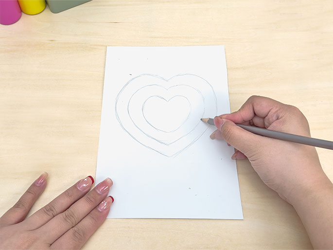 On a plain wooden table, a piece of white paper lies flat. A hand holding a pencil is drawing a series of concentric heart shapes on the paper. Another hand is positioned near the bottom left corner of the paper. The image captures the process of creating a layered heart design.