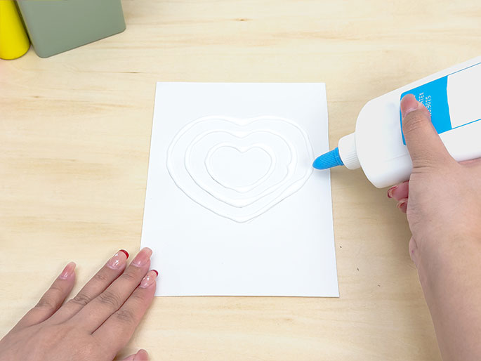 A hand holding a bottle of white glue is applying the glue along the lines of a concentric heart design drawn on a white piece of paper. The paper rests on a light wooden surface. Another hand is placed near the bottom left corner of the paper, seemingly holding it in place. The image depicts the process of preparing for a craft activity, likely involving applying colored material to the glue.