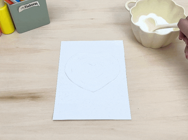 A piece of white paper with a concentric heart design made of glue lies on a light wooden table. The glue appears wet. A pale yellow bowl containing a white granular substance, likely salt or sugar, sits next to the paper. A hand holding a white spoon is taking some of the substance from the bowl, presumably to sprinkle it onto the glue design. The image suggests a stage in a craft project.