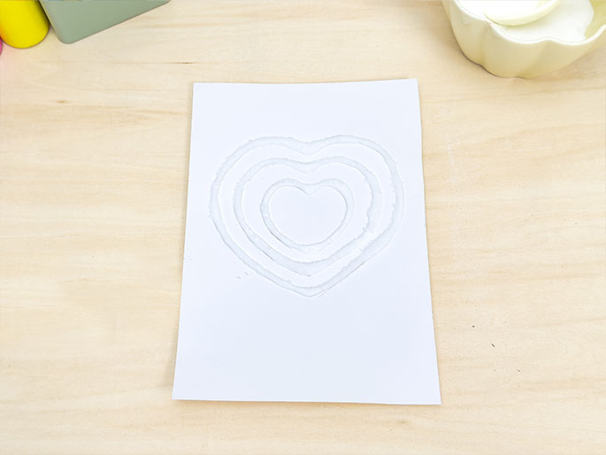 A white sheet of paper on a light wood surface displays a series of concentric heart shapes. The hearts are formed by white granules, likely salt or sugar, adhered to glue. A partially visible pale yellow bowl sits in the upper right corner. The image showcases a simple art or craft project.