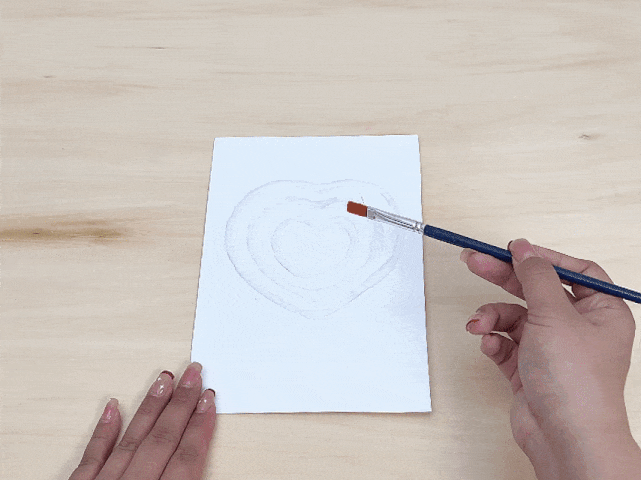 On a light wooden surface, a hand holds a paintbrush with orange paint, about to touch a piece of white paper. On the paper, a series of concentric heart shapes are visible, created with a white granular material, likely salt or sugar, glued to the surface. Another hand is positioned at the bottom left of the paper. The image captures the moment before painting the textured heart design.