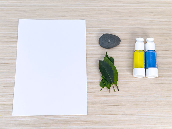 Art supplies are arranged on a light wood-grain table. A blank sheet of white paper lies on the left. To the right, a small gray rock, a cluster of green leaves, and two small bottles of paint (one yellow and one blue) are arranged in a vertical line. The scene appears set for a nature-themed art project.