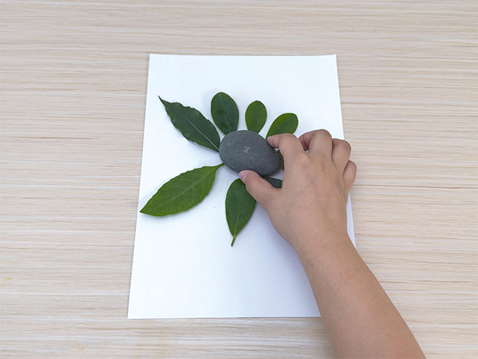 A hand positions a smooth gray rock in the center of a flower-like arrangement of green leaves on a white piece of paper. The paper lies on a light wood-grain table. The leaves, varying slightly in size, are arranged with their stems pointing inwards towards the rock. The hand is placing the rock on top of the leaf stems, completing the nature-inspired design.