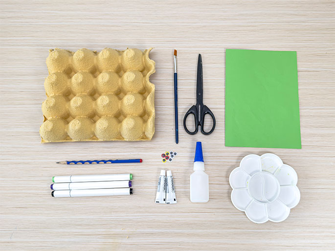 An assortment of craft supplies arranged on a light wood-grain table suggests preparation for a children's art project. The supplies include an empty egg carton, a paintbrush, scissors, a sheet of bright green construction paper, a pencil, several colored markers, small adhesive googly eyes, two tubes of paint (likely acrylic or tempera), a bottle of white glue, and a flower-shaped plastic paint palette with some dried paint remnants. This flat lay image likely illustrates the materials needed for a specific craft activity.