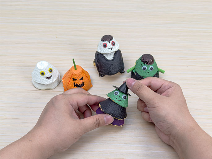 Two hands are placing a small black witch's hat on a Halloween character made from painted egg carton cups. The character has a green face, googly eyes, a black body, and a purple base. Four other egg carton figures are displayed on the light wood-grain table: a ghost, a jack-o'-lantern with a green stem, a vampire, and Frankenstein's monster with green arms. This image demonstrates the completion of a Halloween craft project using recycled materials.