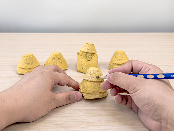 Two hands are shown working on small figures made from egg carton cups on a light wood-grain table. One hand holds a figure while the other draws a face onto it with a pencil. Several other egg carton figures, also with pencil-drawn faces, are visible. This image captures the process of creating simple characters from recycled materials, likely as part of a children's craft activity.