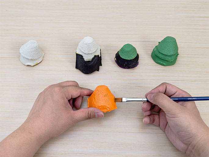 Two hands are painting an orange egg carton cup on a light wood-grain table. A paintbrush applies orange paint to the cup. Several other painted egg carton cups are visible in the background: one unpainted, one with a black base and white top, one with a purple base and green top, and one fully painted green. This image demonstrates the stages of painting upcycled egg carton crafts.
