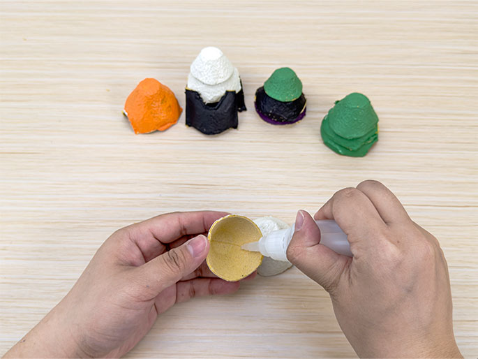 Two hands are applying glue to an egg carton cup on a light wood-grain table. One hand holds the cup while the other squeezes glue from a small bottle. In the background, four other egg carton cups are visible, each painted in different colors or stages of painting (orange, black and white, purple and green, and fully green). This image illustrates a step in a crafting process using recycled materials.