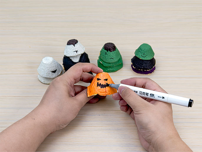 Two hands are drawing a jack-o'-lantern face on an orange painted egg carton cup using a black marker. Several other decorated egg carton figures are lined up on the light wood-grain table behind it. These figures include an unpainted cup, a vampire with a white top and black body, Frankenstein's monster with a green body and brown hair, and another figure with a black body and green top. This image shows the final steps in creating Halloween-themed characters from recycled egg cartons.