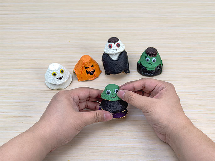 Two hands are adding googly eyes to a small Halloween character made from a painted egg carton cup. The character has a green head, black body, and purple base. Four other completed egg carton figures are arranged on the light wood-grain table: a ghost (white with googly eyes), a jack-o'-lantern (orange with a drawn face), a vampire (white and black with red eyes), and Frankenstein's monster (green with brown hair and googly eyes). This image showcases finished Halloween crafts made from recycled egg cartons.