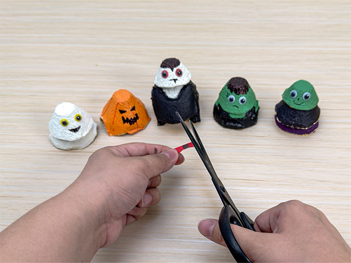 Two hands are cutting a small piece of red and black striped paper with scissors. In the background, five completed Halloween characters made from painted egg carton cups are displayed on a light wood-grain table. The characters are a ghost, a jack-o'-lantern, a vampire, Frankenstein's monster, and a green-faced monster with a black body and purple base. This image suggests an additional crafting step, perhaps creating accessories for the egg carton figures.