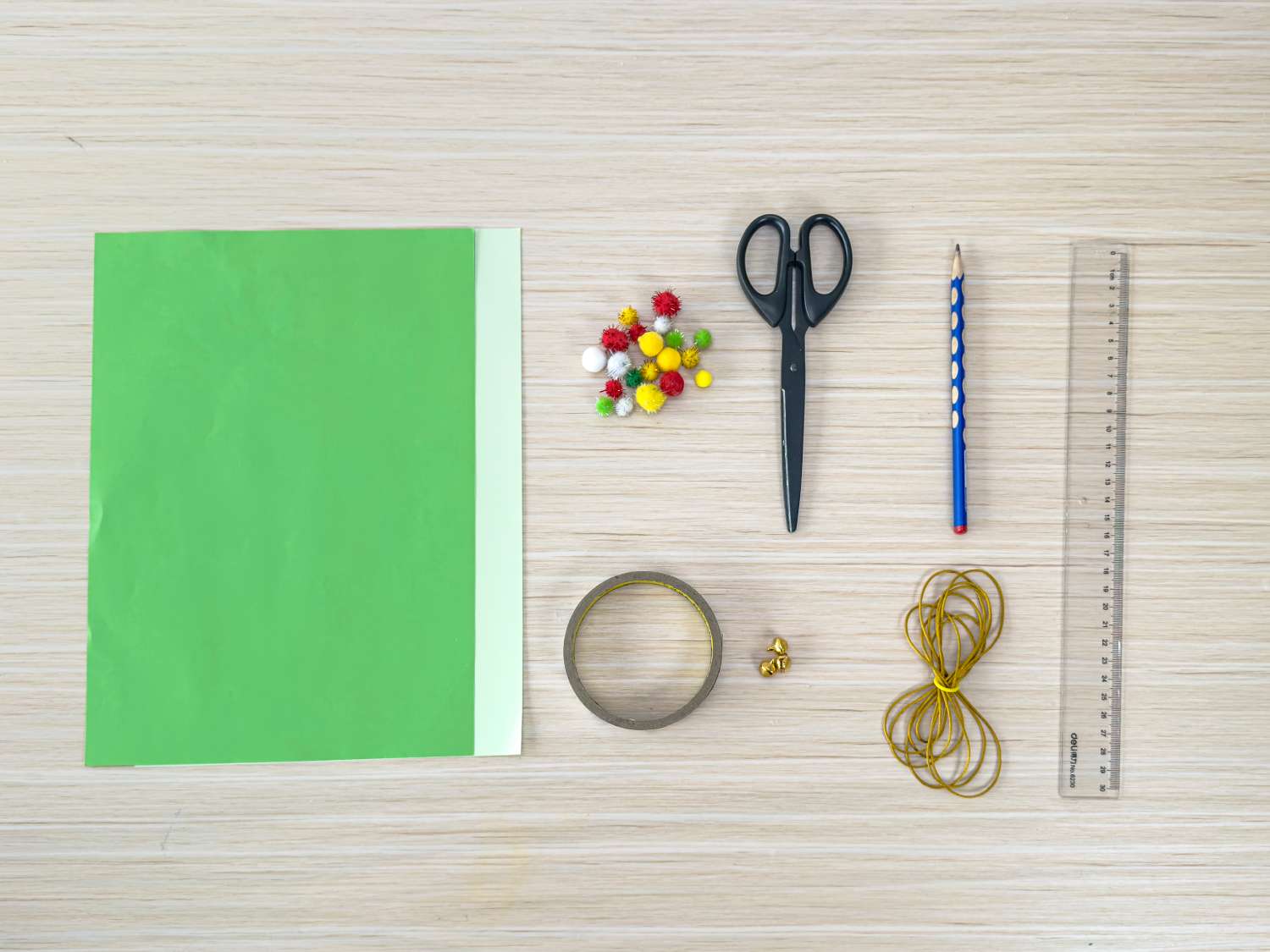 A flat lay showing craft supplies including green paper, pom-poms, scissors, a pencil, a ruler, tape, and string, arranged on a light wood surface.