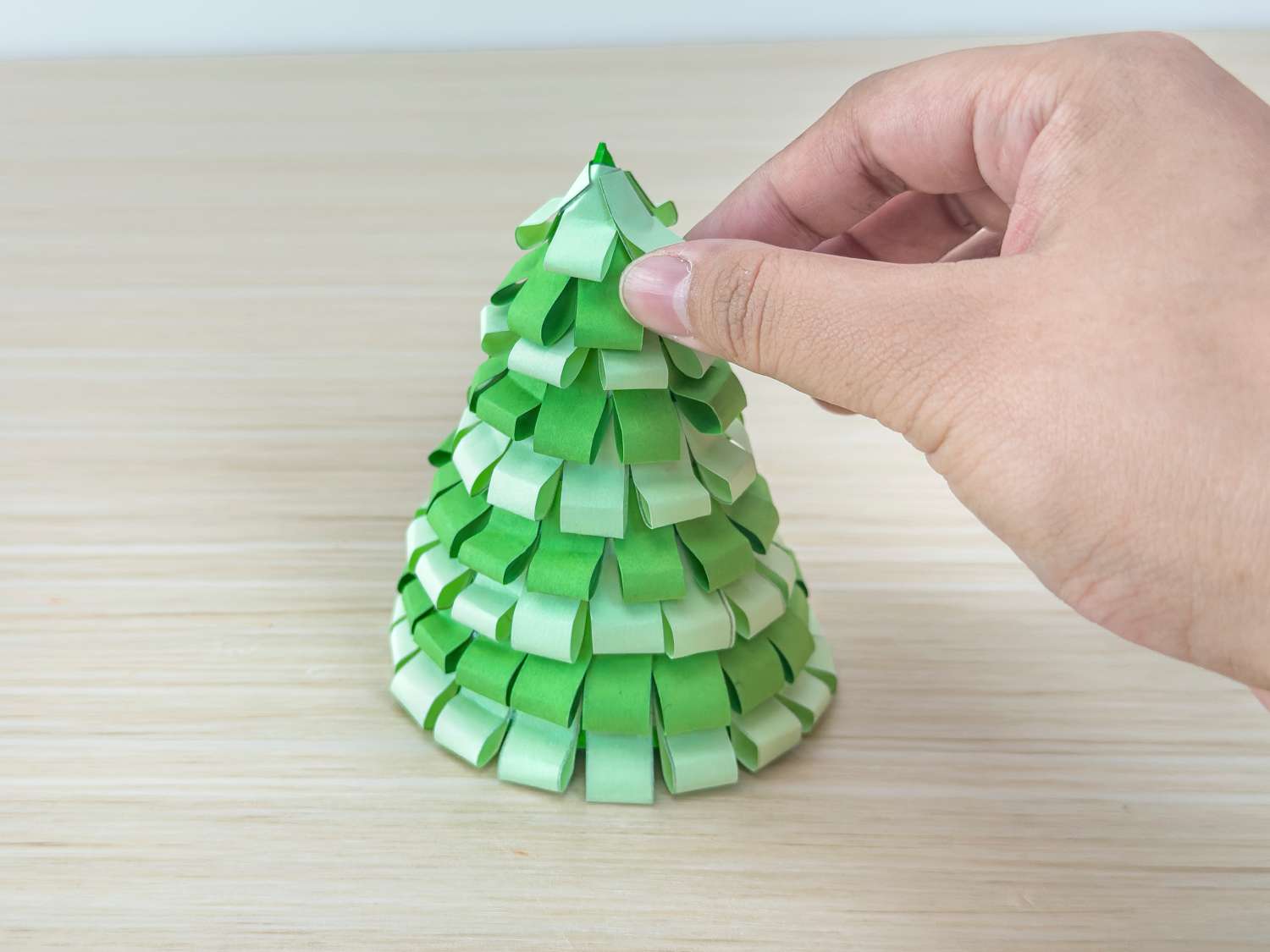 A hand carefully adjusts the top of a small paper Christmas tree made from folded green and pale green paper strips.