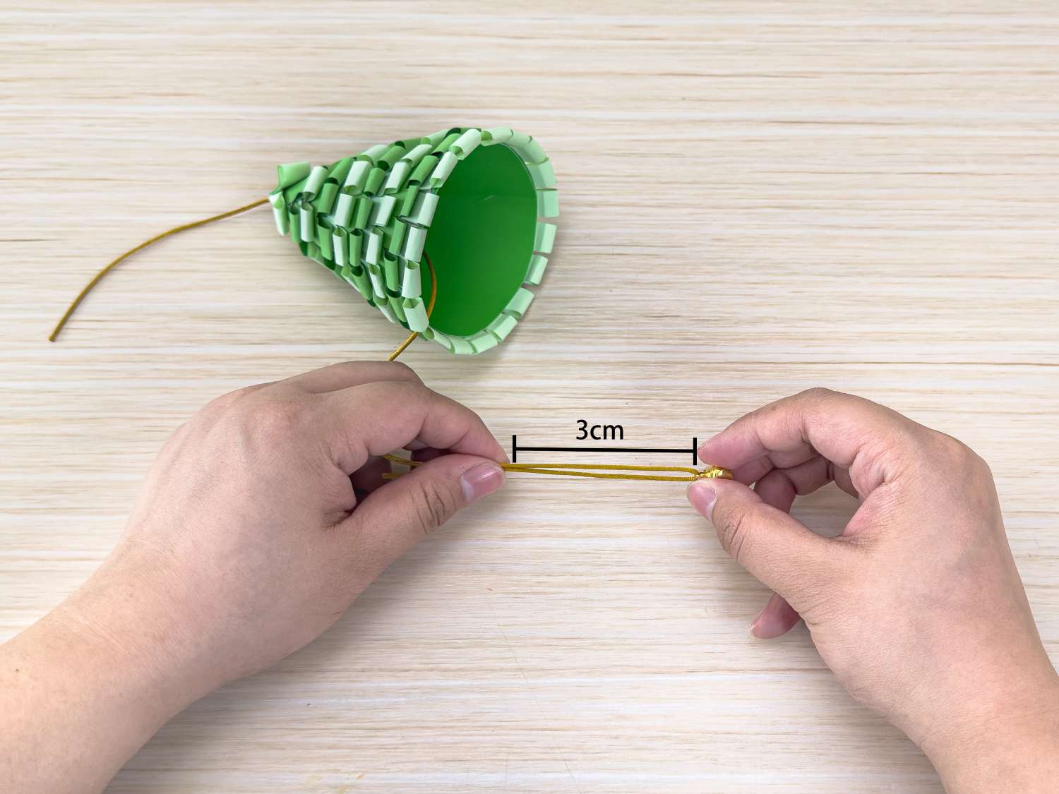 Two hands hold a length of gold cord, measuring 3cm, which is being attached to the top of a paper Christmas tree.