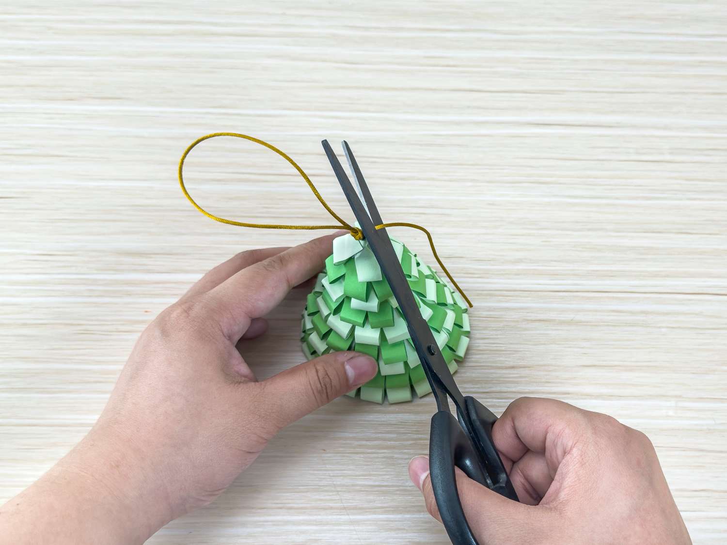 A person uses scissors to trim the excess gold cord from a paper Christmas tree decoration.