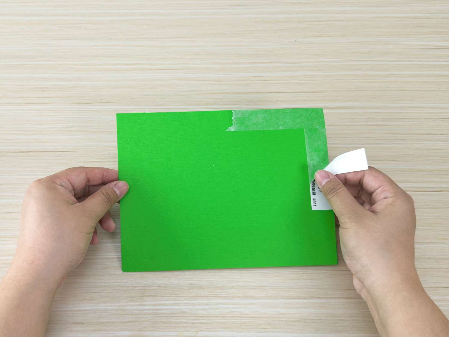 A pair of hands carefully peeling a piece of masking tape from a sheet of bright green paper.