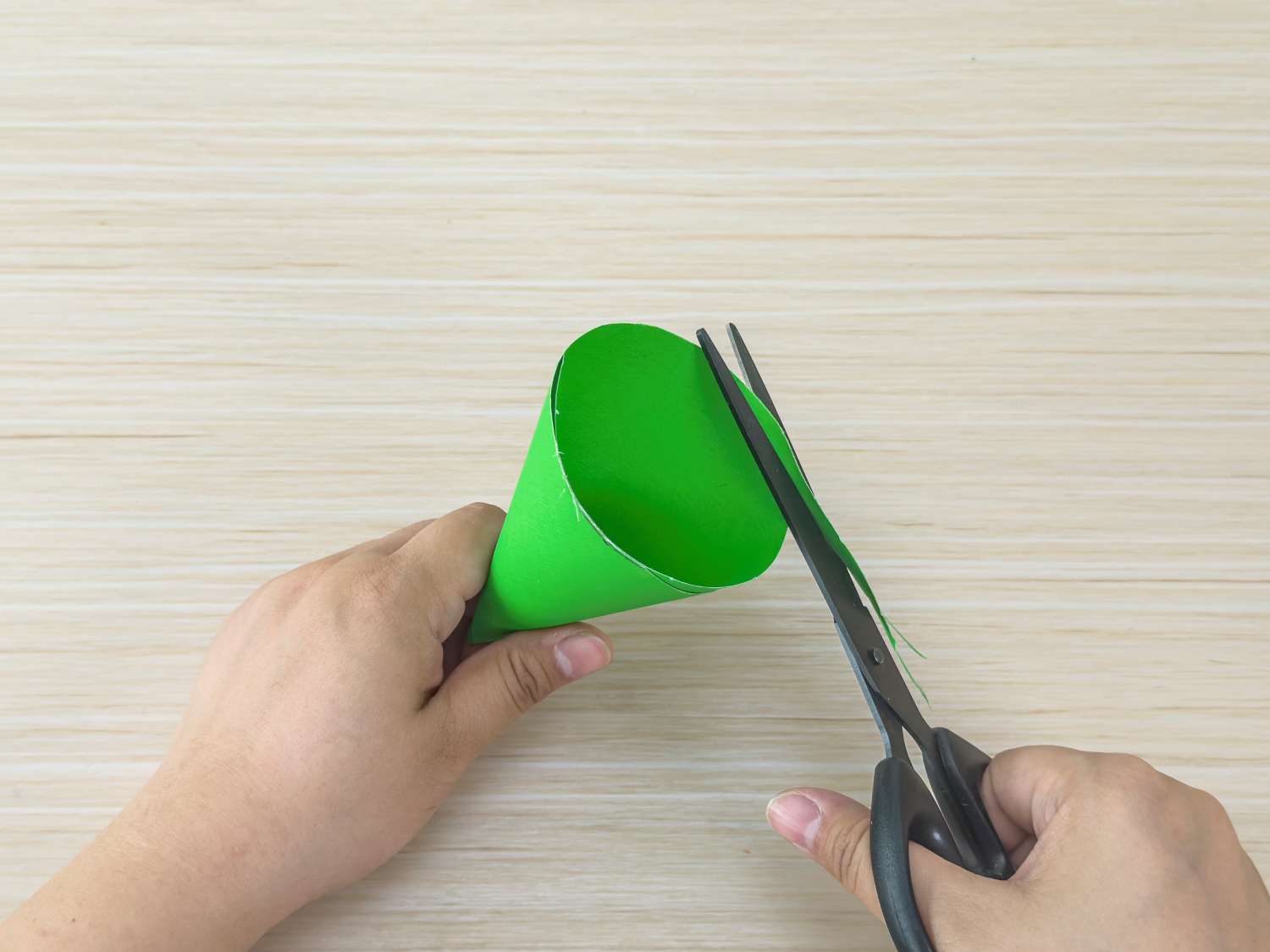 A person uses scissors to trim the excess paper from a green paper cone.