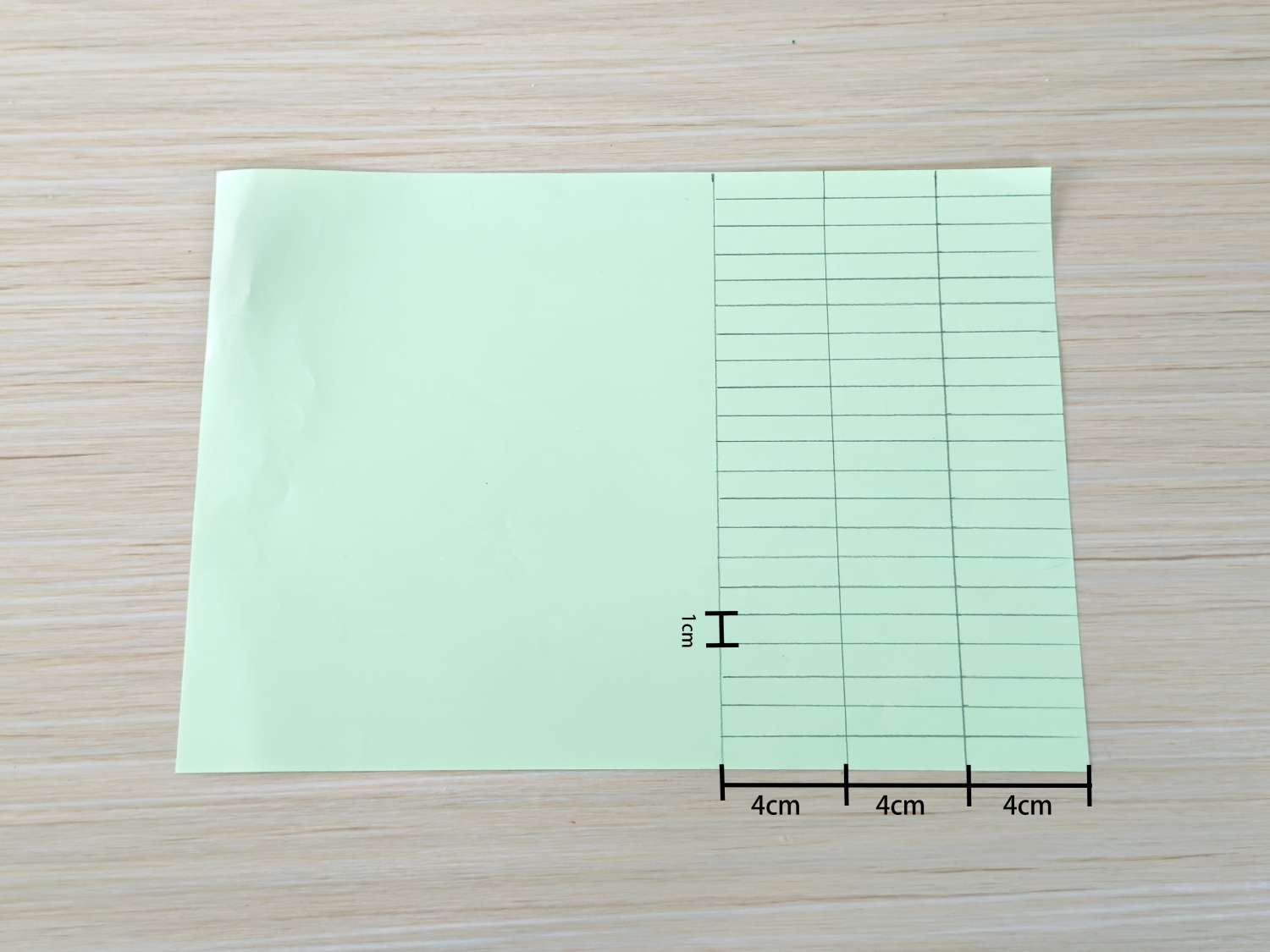A pale green sheet of paper is shown with a grid drawn on one half, measuring 1cm x 12cm, with the other half blank, all on a light wood surface.