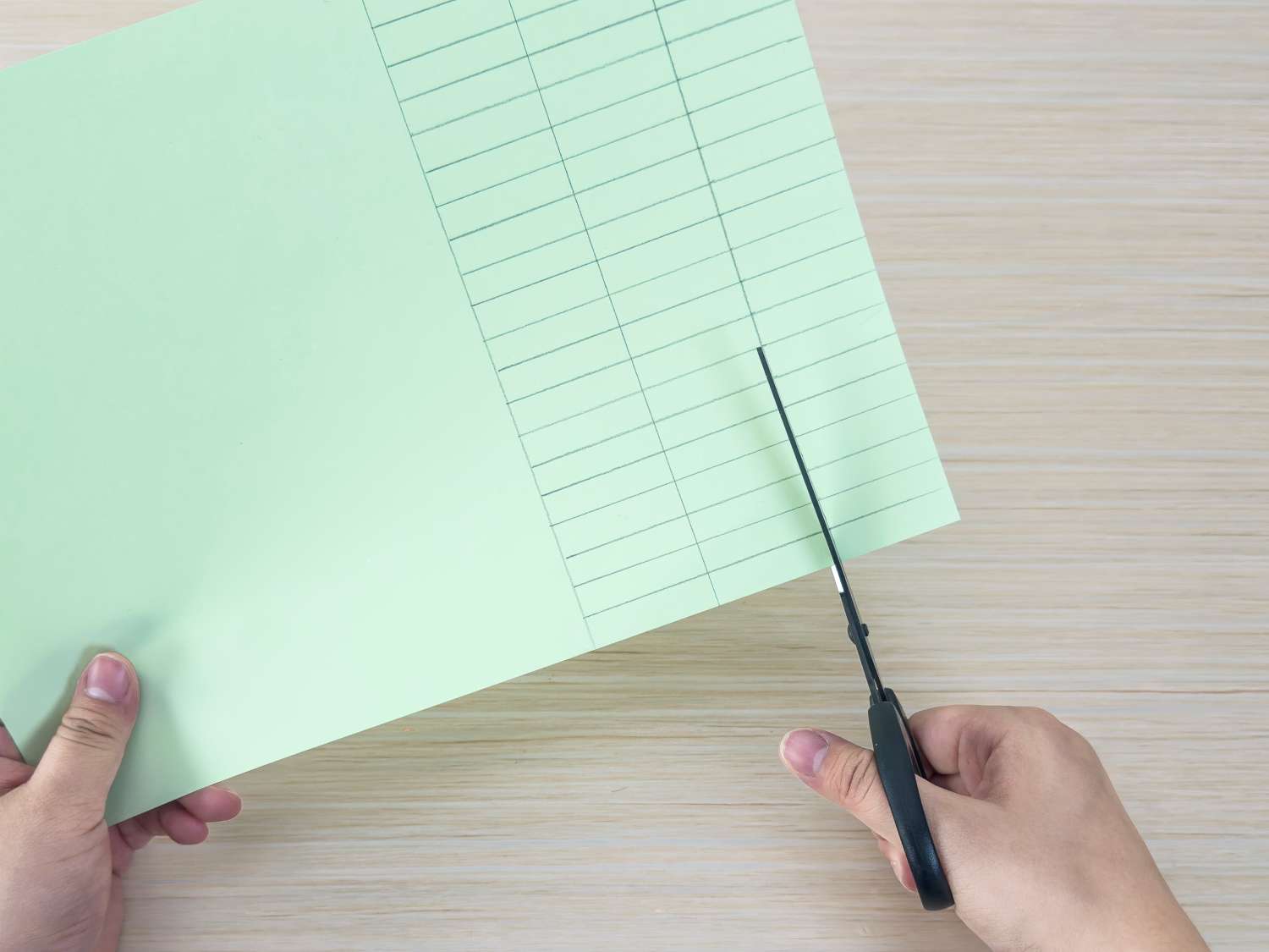 A person uses scissors to cut a pale green sheet of paper that has a grid drawn on one half.