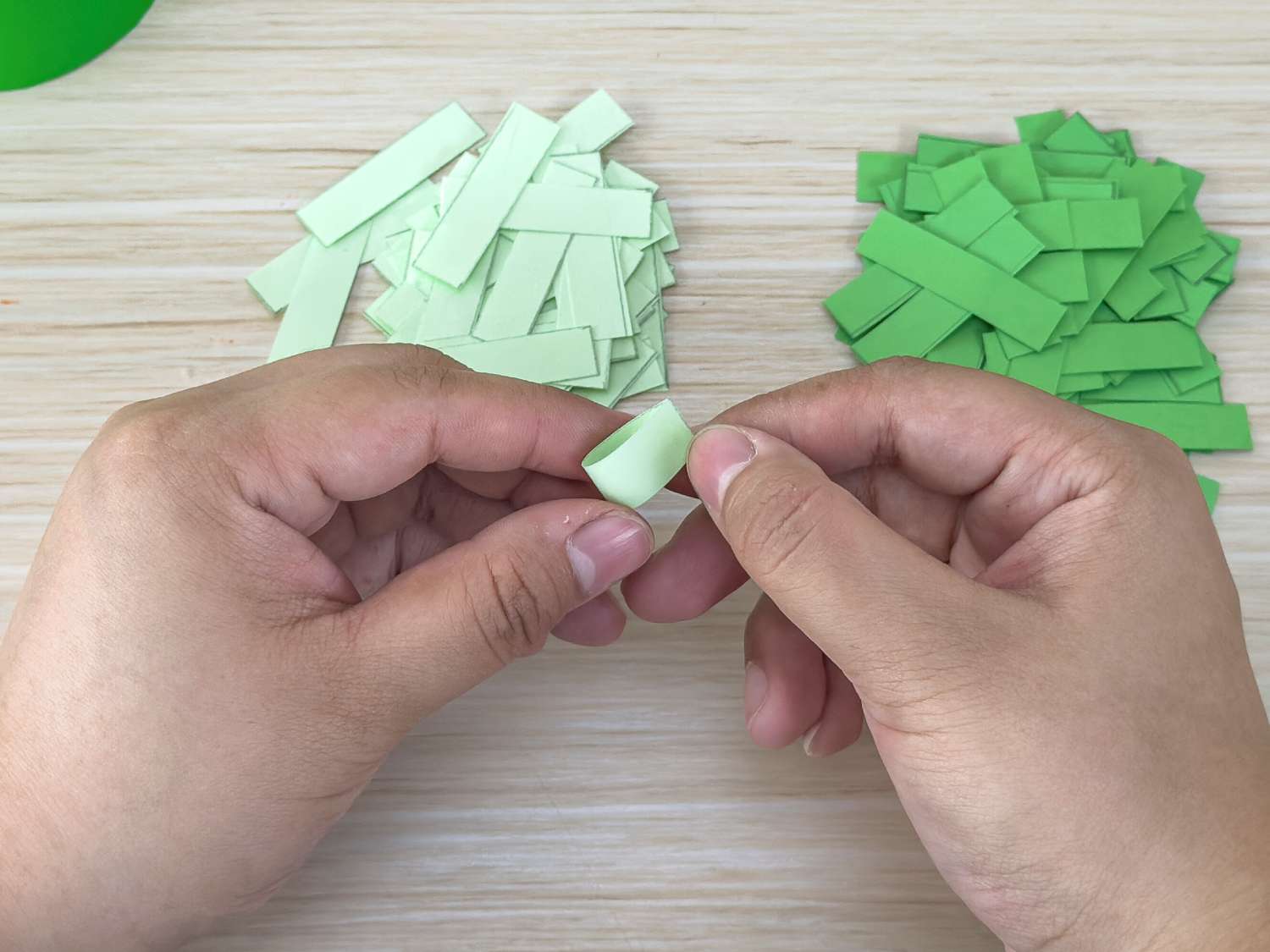 A person's hands are shown rolling a small strip of pale green paper into a tiny tube, with piles of cut paper strips in two shades of green nearby.