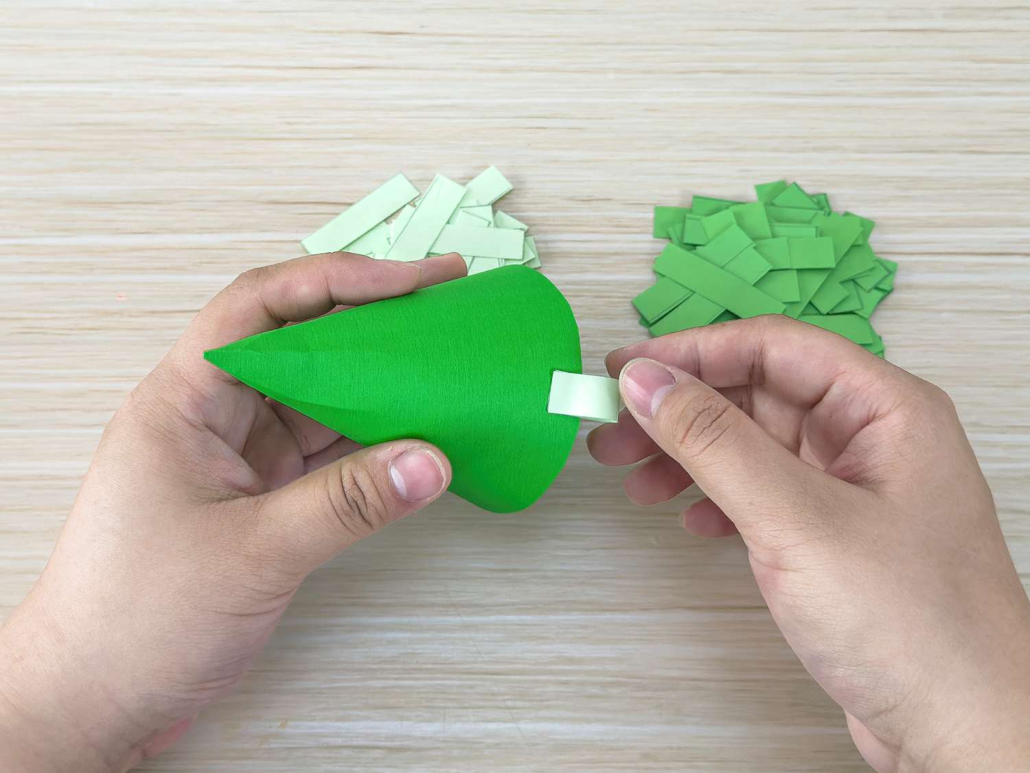 A person is shown securing a small rolled strip of pale green paper to the top of a green paper cone, with piles of cut paper strips in two shades of green nearby.