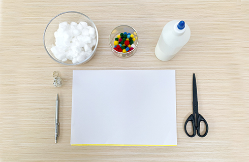 Gather your materials for some festive crafting! A bowl of cotton balls, colourful pom-poms, glue, a piece of paper, a pencil, and scissors are arranged on a light wood surface, ready to create a magical Christmas tree night light. Let's get making!