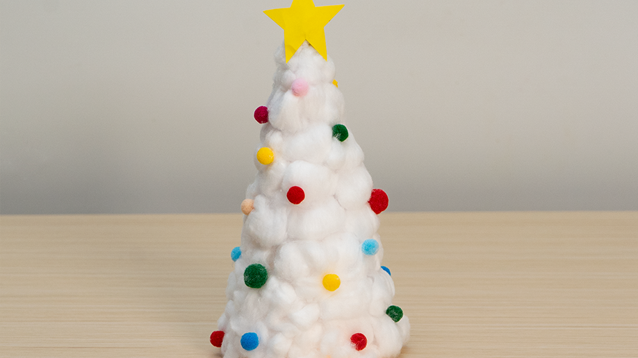 A delightful miniature Christmas tree, crafted from cotton balls and brightly coloured pom-poms, sits proudly on a light wood surface, topped with a cheerful yellow star. Ready to spread some festive cheer!