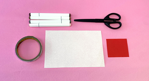 Gather your supplies for some festive crafting fun! A selection of coloured markers, scissors, double-sided tape, a sheet of white paper, and a small square of red paper are neatly arranged on a pink surface, ready to create some dancing reindeer. Let's get started!