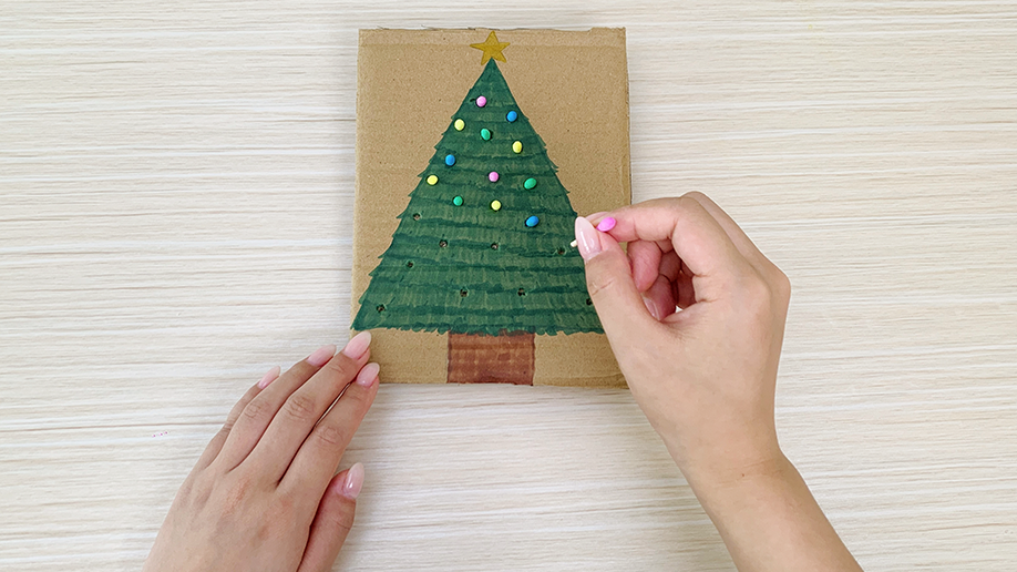 A hand carefully pushes colorful cotton buds into the holes of the painted Christmas tree, adding a festive touch. It's all looking rather jolly, wouldn't you say?