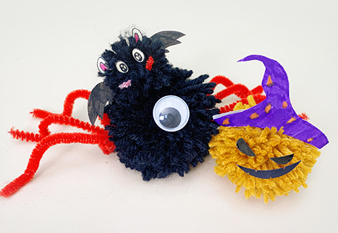 Two charming Halloween-themed pom-poms are displayed on a white surface. One is a black pom-pom representing a bat, complete with small paper wings and googly eyes, attached to bright red pipe cleaner legs forming a spider. Beside it sits a yellow pom-pom depicting a jack-o'-lantern, adorned with a purple paper witch's hat and simple black features. The bright colours and whimsical designs create a delightful and festive scene, suggesting handmade Halloween decorations.