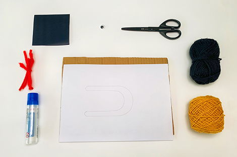 The image displays various craft materials arranged on a white surface, ready for a project. These include balls of black and yellow yarn, a pair of scissors, a small bottle of adhesive, a piece of red pipe cleaner, a small black square of card, a single googly eye, and a piece of cardboard supporting a sheet of white paper with a drawn outline resembling a large, rounded 'U' shape. The items are neatly positioned, suggesting preparation for a structured craft activity, possibly involving pom-pom making.